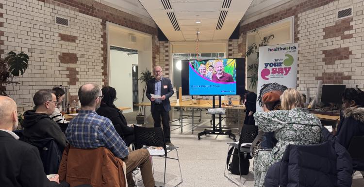 A speaker presenting to a seated audience in a modern, tiled space with a Healthwatch banner visible.