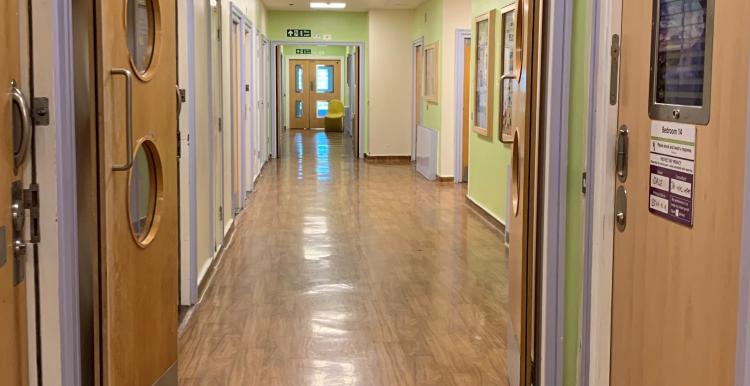 A brightly lit hospital corridor with light green walls and wooden flooring. The hallway features several doors on both sides, some with windows, and a fire exit sign above leading forward. The far end of the corridor is visible, with a door slightly ajar and a chair in the distance.