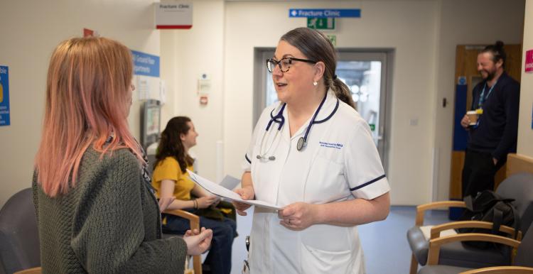 A healthcare professional in a white uniform and stethoscope speaks with a woman with pink hair in a hospital waiting area. Behind them, a young woman in a yellow shirt is seated, and a man stands in the background holding a cup. The environment features signage for a fracture clinic and chairs for waiting patients.