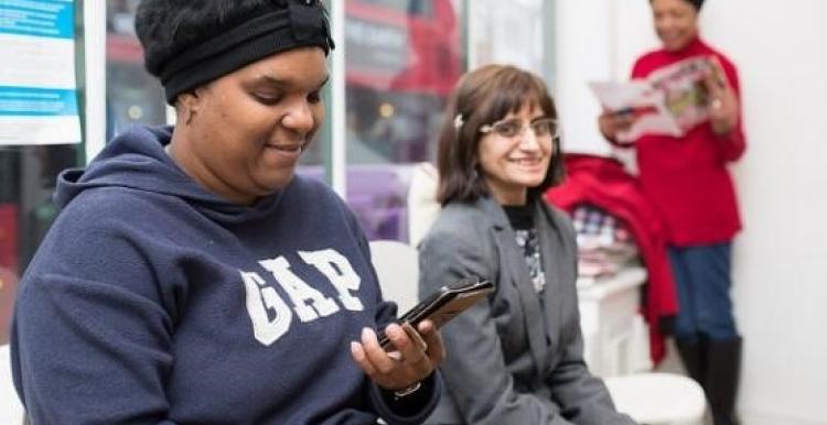 Three women in a bright room; one in a navy hoodie smiles at her phone, another in a gray blazer smiles at the camera, and a third in a red top reads a magazine in the background. A double-decker bus is visible through the window.