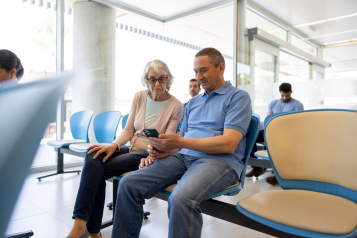 Two people sitting in a waiting room, looking at a smartphone together. An older woman wearing glasses and a light pink cardigan watches as the man beside her, dressed in a light blue polo shirt, holds the phone. Other people are seated in the background.