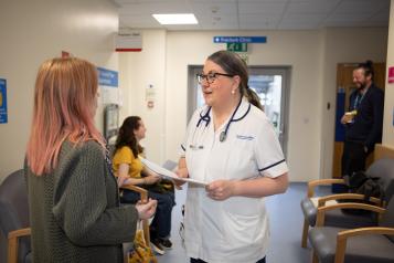 A healthcare professional in a white uniform and stethoscope speaks with a woman with pink hair in a hospital waiting area. Behind them, a young woman in a yellow shirt is seated, and a man stands in the background holding a cup. The environment features signage for a fracture clinic and chairs for waiting patients.