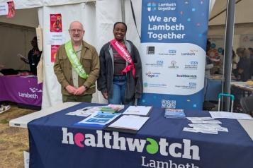 A man and a woman smiling and standing behind a table that says Healthwatch Lambeth