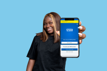 Woman smiling holding a phone towards the camera. On her screen is the NHS App log-in screen.