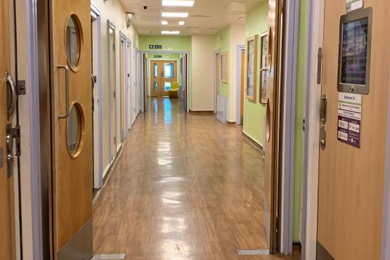 A brightly lit hospital corridor with light green walls and wooden flooring. The hallway features several doors on both sides, some with windows, and a fire exit sign above leading forward. The far end of the corridor is visible, with a door slightly ajar and a chair in the distance.