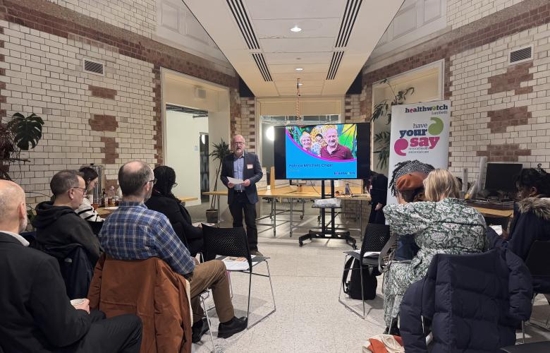A speaker presenting to a seated audience in a modern, tiled space with a Healthwatch banner visible.