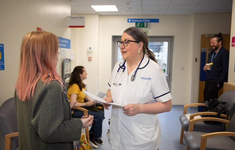 A healthcare professional in a white uniform and stethoscope speaks with a woman with pink hair in a hospital waiting area. Behind them, a young woman in a yellow shirt is seated, and a man stands in the background holding a cup. The environment features signage for a fracture clinic and chairs for waiting patients.