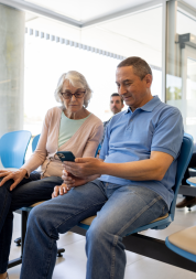 Two people sitting in a waiting room, looking at a smartphone together. An older woman wearing glasses and a light pink cardigan watches as the man beside her, dressed in a light blue polo shirt, holds the phone. Other people are seated in the background.