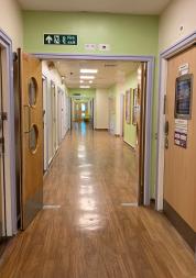 A brightly lit hospital corridor with light green walls and wooden flooring. The hallway features several doors on both sides, some with windows, and a fire exit sign above leading forward. The far end of the corridor is visible, with a door slightly ajar and a chair in the distance.