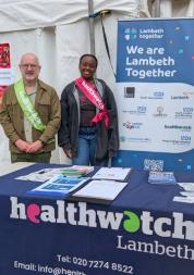 A man and a woman smiling and standing behind a table that says Healthwatch Lambeth
